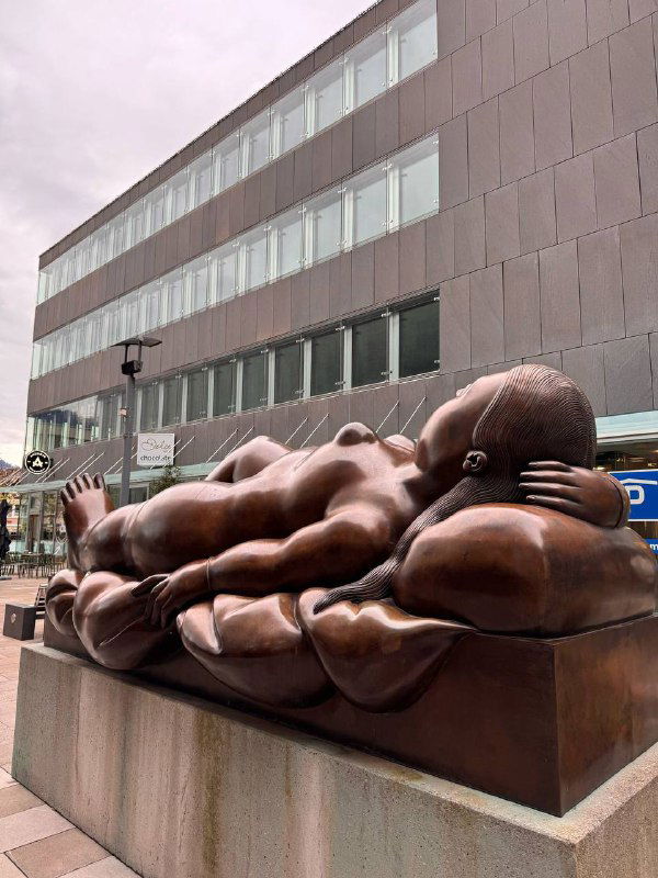 Крупная скульптура лежащих фигур перед современным зданием в городском пространстве — художественный объект у городской площади.