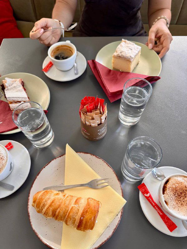 Стол в кафе с чашками кофе, тарелками с трубочками и кусочками пирожных, руки гостей и стаканы воды в кадре.