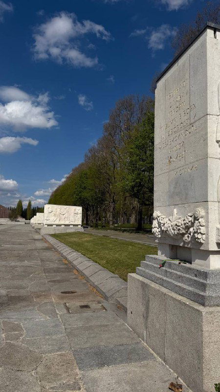 Фрагменты монументальных плит и барельефов мемориального комплекса, аккуратные каменные облицовки и зелёная полоса рядом с проходом.