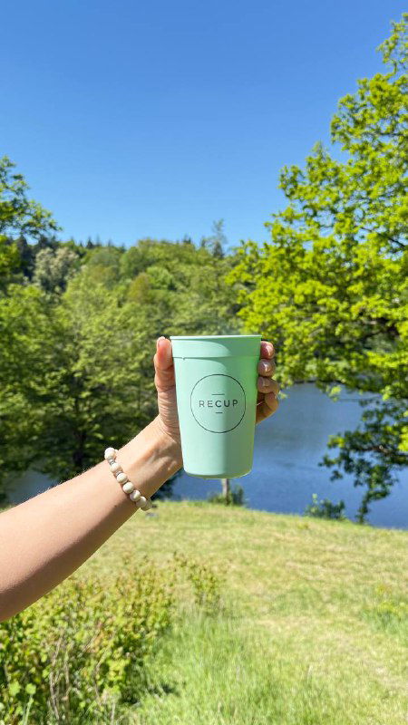 Рука с браслетом держит светло-зелёную чашку RECUP на фоне реки и леса — летний пейзаж, чашка остаётся в фокусе.
