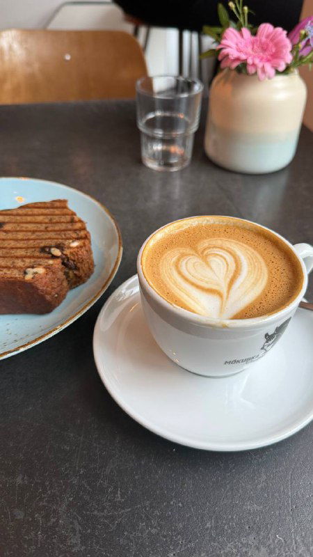 Капучино с латте-артом в белой чашке и кусок торта на тарелке на столе в кафе, рядом ваза с цветами и стеклянный стакан.