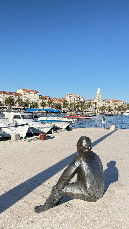 Прибрежная набережная с причаленными лодками и скульптурой на причале; вдалеке виден исторический город и колокольня на фоне голубого неба.