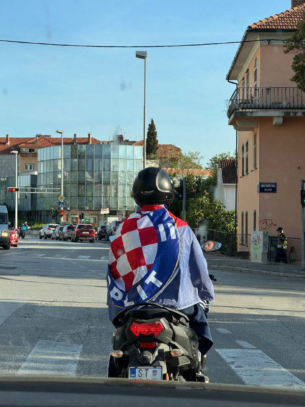 Мотоциклист в городской улице с накидкой в узор хорватского флага: движение по узкой дороге среди жилых зданий в небольшом городке.