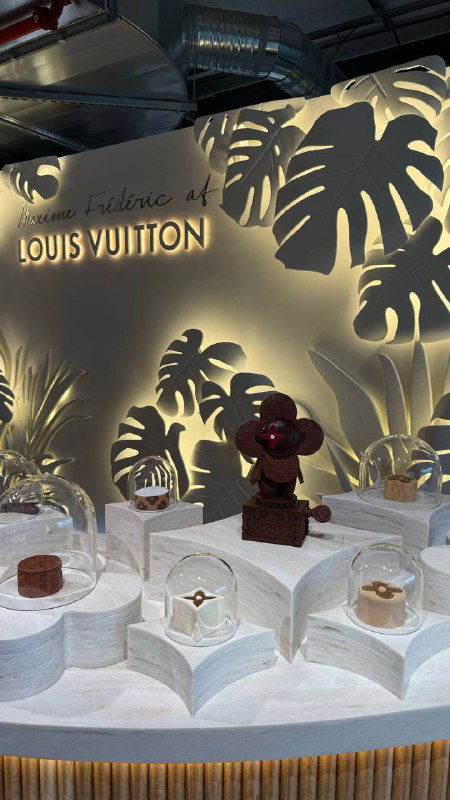 Витрина Louis Vuitton: декоративные объекты, шоколад и мерч на подиуме с подсветкой и логотипом бренда в зоне выставки.