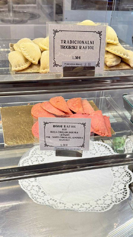 Витрина кондитерской с подносом розовых и бежевых сладких равиоли, бумажные таблички с названиями и ценой видно через стекло.