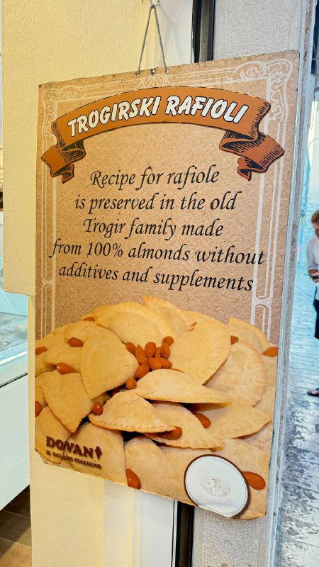 Рекламный плакат с надписью "Trogirski RafIoli" и описанием семейного рецепта в витрине кафе, изображены золотистые равиоли.