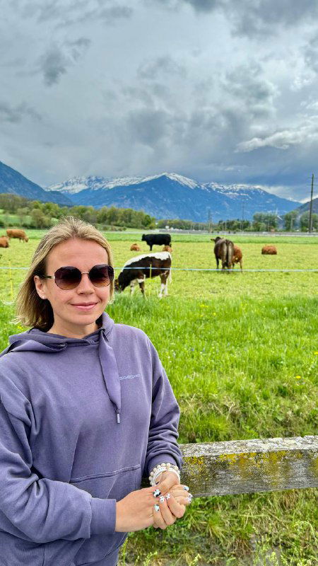 Автор на переднем плане в солнцезащитных очках, за ней зелёное поле с пасущимися коровами и снежными вершинами альпийских гор на горизонте.