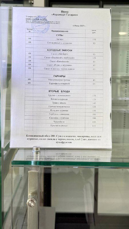 Фотография бумажного меню или прайса в стеклянной витрине с перечнем блюд и цен, видимы позиции столовой и стоимость некоторых блюд.