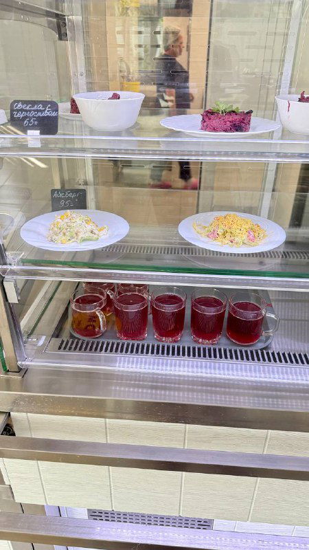 Витрина с порционными блюдами и компотом: тарелки с салатами, горячими блюдами и ряд баночек с компотом на прилавке столовой.