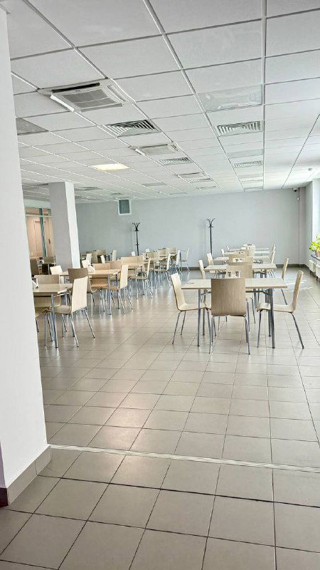 Пустой просторный зал столовой с однородной мебелью, столы и стулья расставлены в простом столовском стиле, видна светлая плиточная отделка.