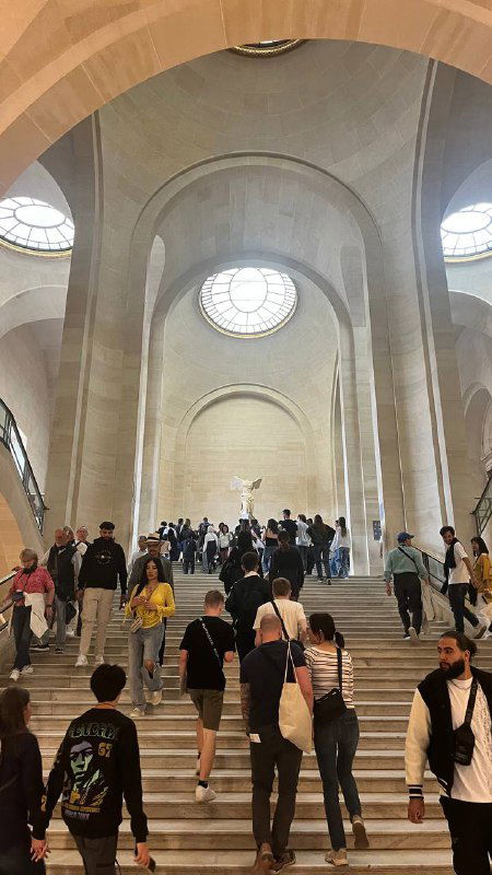 Широкая лестница в интерьере Лувра с арочными сводами и круглыми окнами: посетители поднимаются по ступеням в музейном пространстве