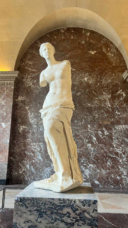 Статуя Венеры Милосской в Лувре на мраморной подставке: классическая скульптура без кистей рук на фоне тёмной стены и пола