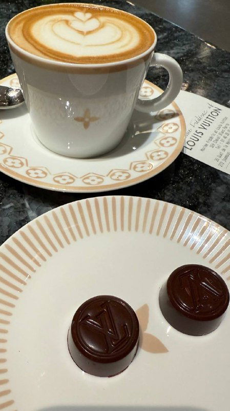 Кофе и шоколад в кафе Louis Vuitton: чашка капучино с латте-арт и две круглые шоколадные конфеты на белой тарелке рядом с меню