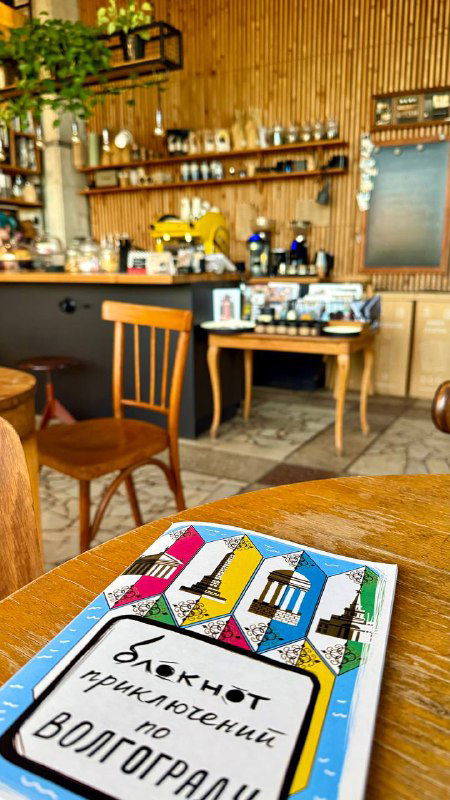 Крупный план стола с блокнотом «Блокнот путешественника по Волгограду» на фоне интерьера кофейни и деревянной мебели.