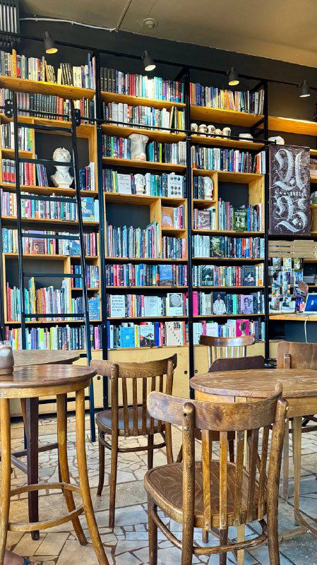 Стеллажи с книгами в кофейне внутри библиотеки: деревянные полки, много изданий и свободные столики для чтения.