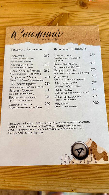 Меню кофейни с перечнем напитков и ценой, оформленное в книжном стиле и размещённое на деревянном столе внутри заведения.