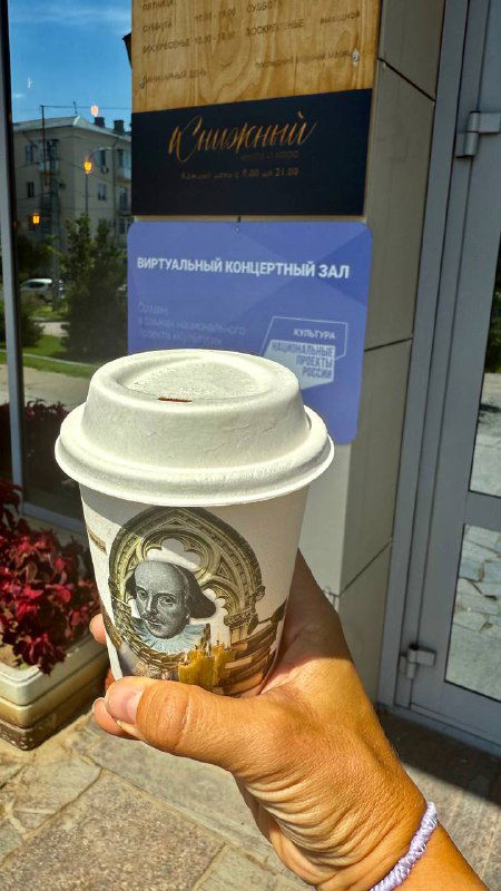 Рука держит бумажный стакан с принтом и крышкой на фоне стеклянной витрины; кадр с кофе навынос у входа в библиотечную кофейню.
