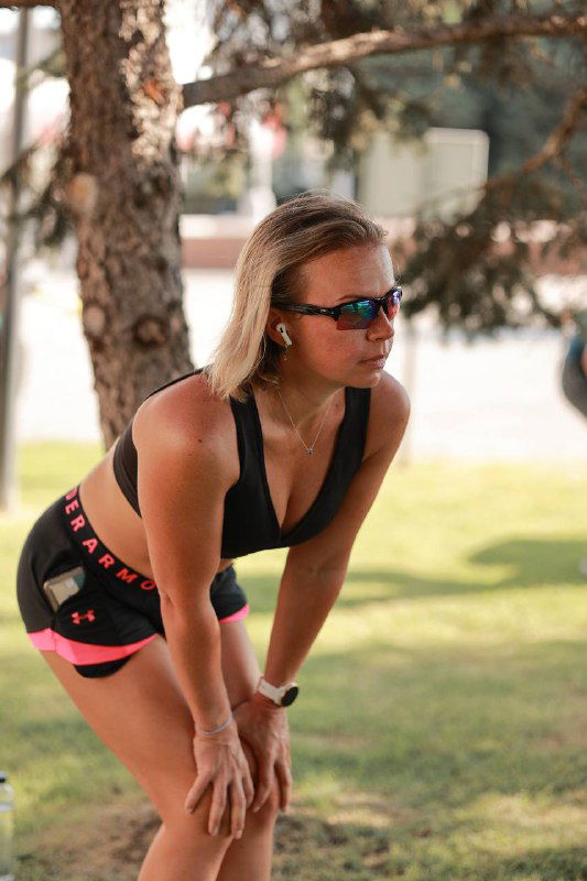 Спортсменка в очках и наушниках отдыхает, опершись о колени в парке; кадр подчёркивает небольшой перерыв после пробежки и городской зелёный фон.