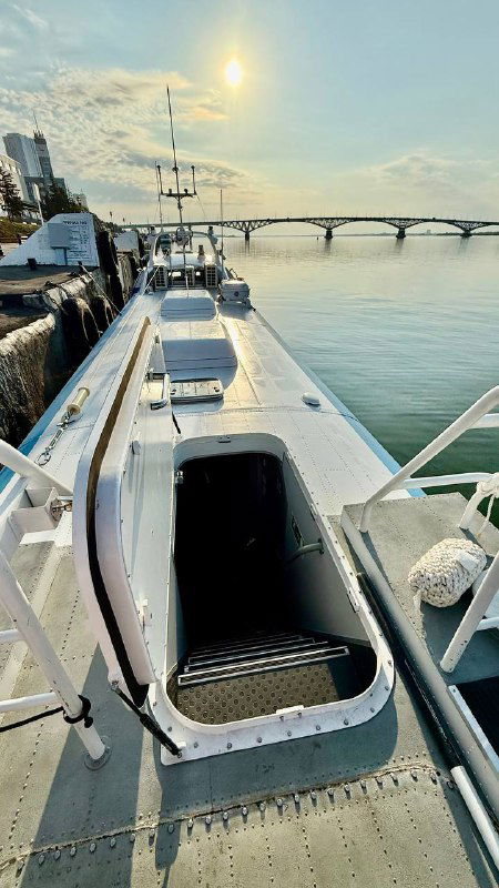Палуба катера с открытой дверью в салон, ступеньки и поручни видны на фоне реки и моста, лёгкое вечернее освещение и река.