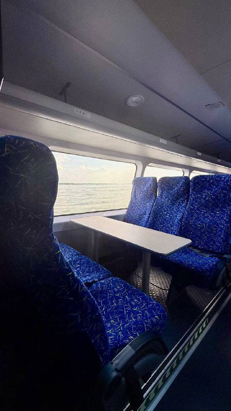 Ряд синих кресел внутри катера, рядом столик у окна, солнечный свет сквозь стекло и вид на Волгу создают атмосферу комфорта в пути.