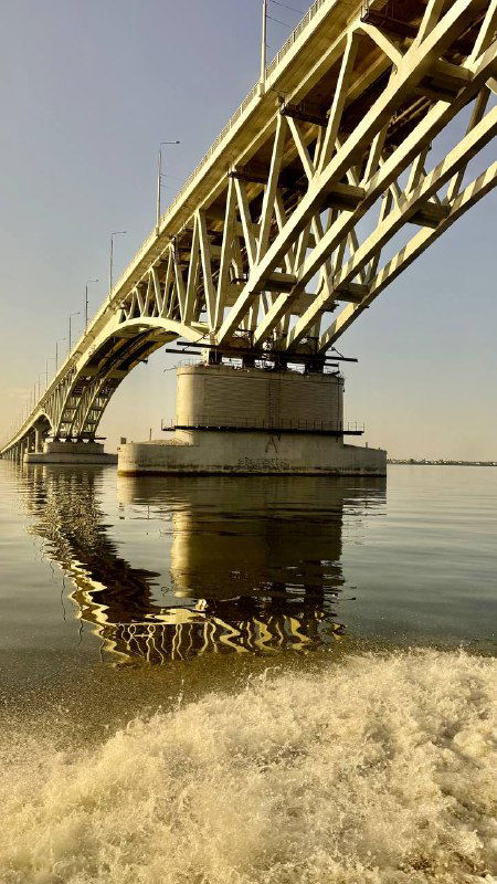 Арочный железный мост через Волгу с массивной опорой и отражением в воде, кадр сделан с кормы катера во время движения по реке.