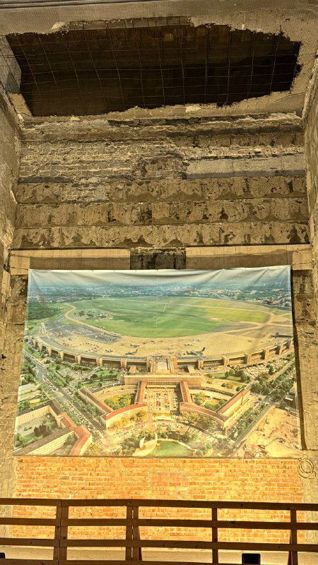 Интерьер заброшенного здания с дырами в потолке и повешенным плакатом с панорамой аэропорта — контраст между разрухой и сохранившейся картиной.