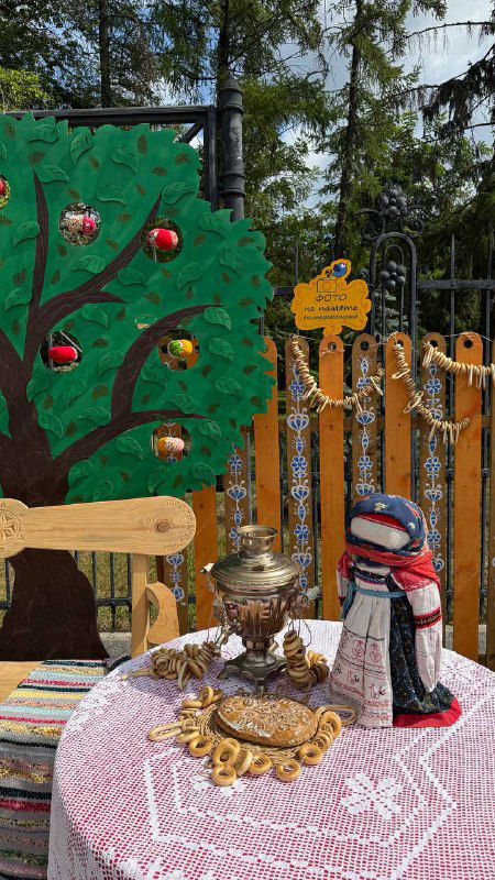 Павильон с хэндмейд-изделиями: самовар, расписные предметы и текстильная кукла на фоне декоративного деревянного забора и ярких украшений.