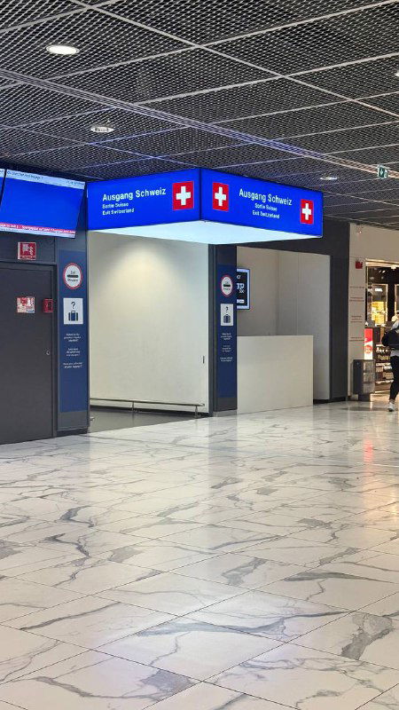 Внутренний зал с указателями выхода в Швейцарию («Ausgang Schweiz») и табличками с швейцарским крестом, пол из светлого мрамора и пустая просторная зона.