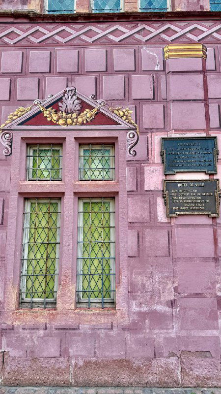 Фрагмент расписанного фасада в розовых тонах с декоративными вставками, оконными рамами и мемориальными табличками на стене дома.