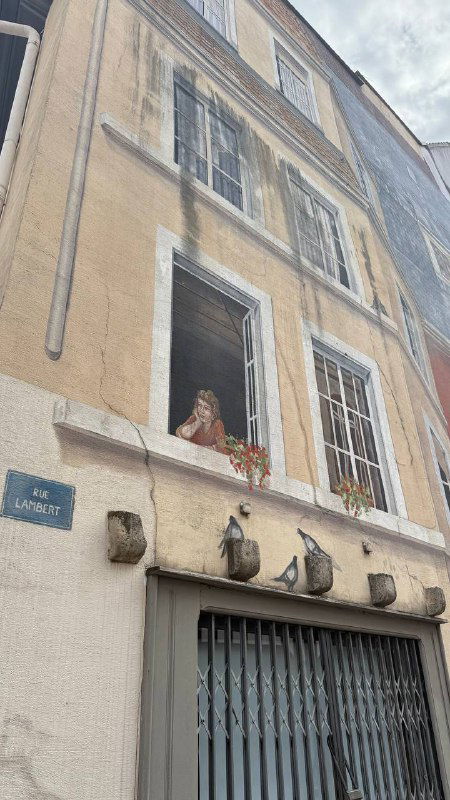 Угол дома с Trompe-l'œil росписью: изображённые архитектурные элементы и окна имитируют глубину и декор на плоской стене здания.