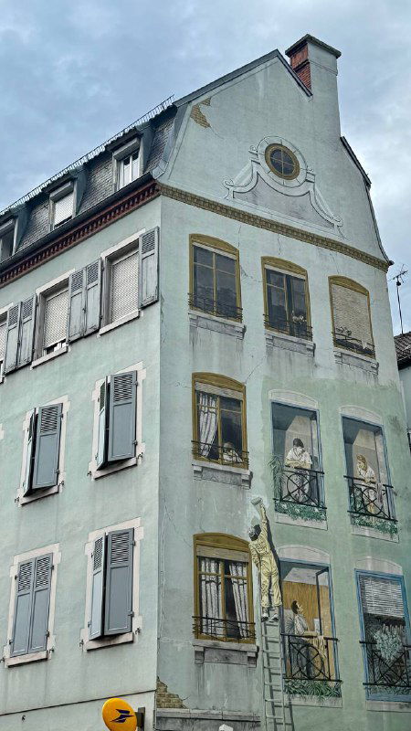 Пастельно-зелёный жилой дом с живописными элементами на фасаде и ставнями на окнах в спокойном квартале Мюлуза под облачным небом.