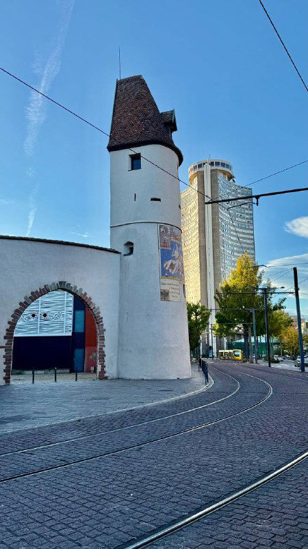 Одна из старых городских башен Мюлуза: белая башня с аркой у основания и трамвайными путями на переднем плане в осеннем свете.