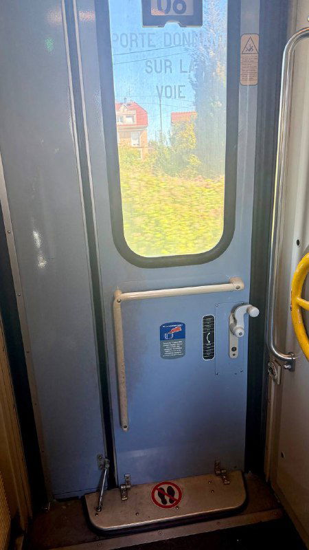 Внутренняя дверь вагона с видом на наружу через стекло: сиденья, поручень, ручка, отражение и солнечный пейзаж за окном.