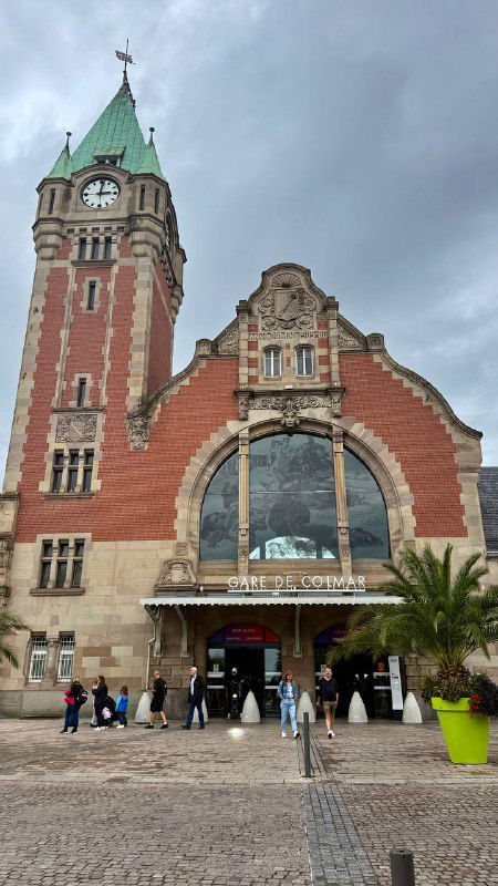 Фасад старого железнодорожного вокзала во Франции с часовой башней, декоративными скульптурами и арочным входом, люди у дверей.