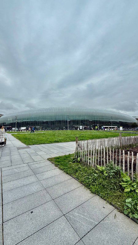 Современный стеклянный вокзал крупным планом: куполообразный фасад из стекла, передняя площадь и урбанистический ландшафт вокруг здания.