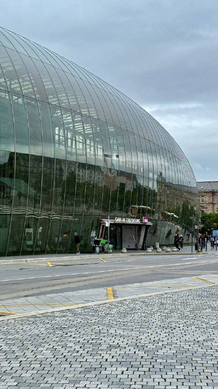 Близкий ракурс стеклянного фасада вокзала с заметным входом, отражениями неба на панелях, пешеходами и городской мебелью на площади.