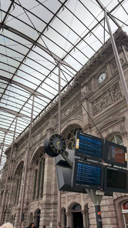 Внутренний зал большого европейского вокзала с массивной стеклянной крышей, настенными часами, электронными табло и движением пассажиров.