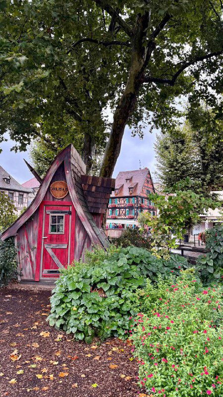 Маленький домик в стиле сказки среди зелени и кустов в парковой зоне Кольмара на фоне облачного неба и старых зданий.