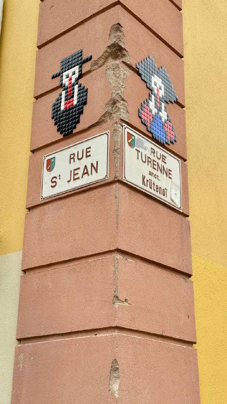 Угловой фасад с уличными табличками Rue St-Jean и Rue Turenne, декоративные мозаики на углу дома и городской исторический антураж.