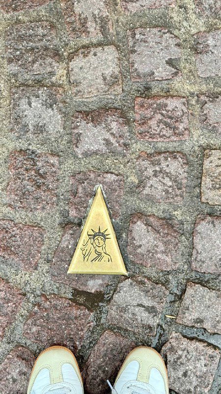 Крупный план мощёной каменной тропинки с небольшим треугольным знаком на земле и сапоги/тапочки человека в кадре, уличная деталь.