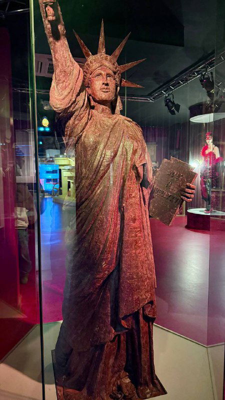 Миниатюрная Статуя Свободы в музее Кольмара на витрине, музейное освещение и экспозиция шоколадных реликвий на заднем плане.