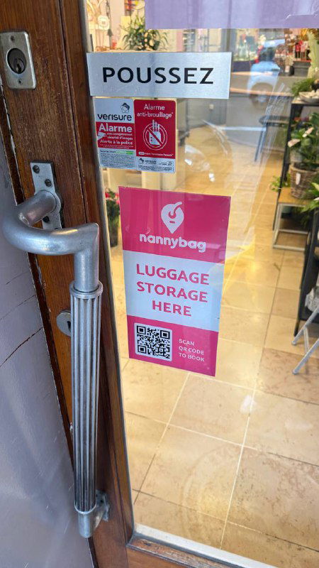 Дверь магазина с наклейками сервиса хранения багажа: розовая наклейка 'LUGGAGE STORAGE HERE', логотип nannybag и QR-код на стекле.