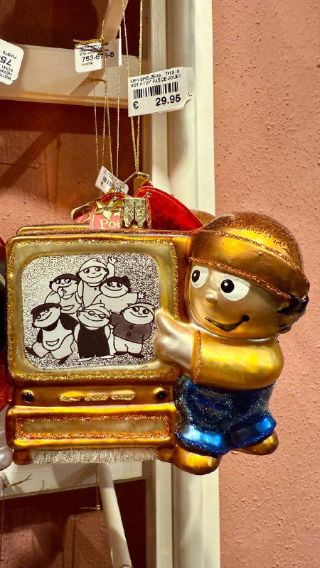 Игрушка в виде человечка с телевизором и выразительным лицом, подвешенная на крючке среди других украшений и ценников в отделе магазина.