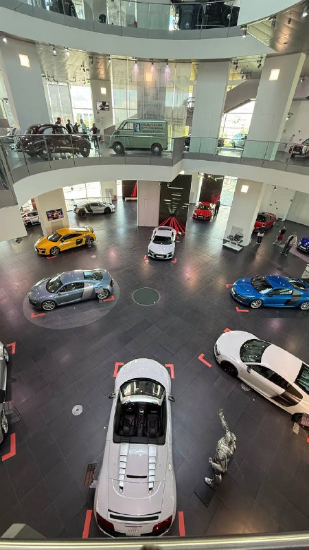 Круглый выставочный зал с экспонатами Audi на полу и автомобилями, расставленными по спирали на нескольких уровнях внутри просторного светлого пространства музея.