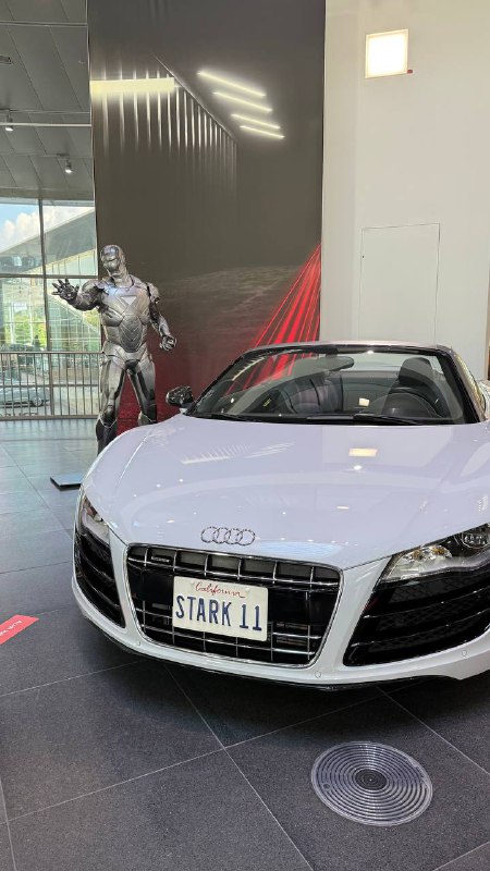 Белый спортивный автомобиль Audi с табличкой STARK II на переднем плане и декоративной фигурой рядом, экспонат из кинопоказательной зоны по мотивам «Железного человека».