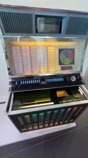 Музыкальный автомат в зоне 70-х в музее Audi: пульт с символикой эпохи и переливающейся панелью выбора мелодий, интерактивный ретро-экспонат на фоне стены.