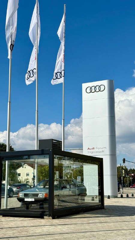 Флаги Audi и компактная остановка/вход рядом с офисным зданием, вид с улицы на логотип бренда и окружающую инфраструктуру у форума Audi в Ингольштадте.