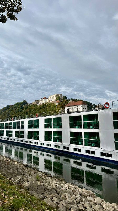 Круизный теплоход у набережной Пассау: белый корпус, окна кают и отражение в спокойной воде рядом с каменной набережной города.