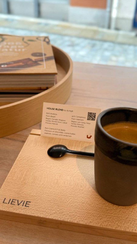 Кофе в чашке на деревянном подносе в кофейне Lievie: карточка зерна, маленькая ложка и аккуратная подача на столе у окна кафе.
