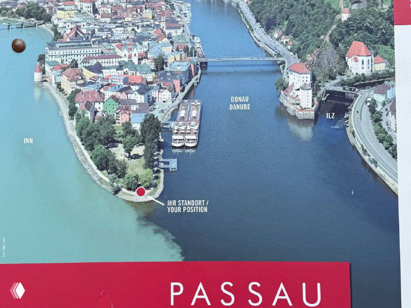 Информационный стенд с картой Пассау: схема слияния рек, отметки маршрутов и контрастная иллюстрация берега с бирюзовой водой.